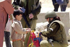 An Israeli soldier in full uniform -- helmut, gun, bullet-proof vest -- examines a small child's backpack.
